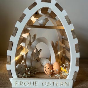 Könnte beinhalten: Weiße, hölzerne Oster-Eier-Dekoration mit einem Vogel, einem Hasen und Eiern im Inneren. Die Dekoration hat ein Licht im Inneren und steht "Frohe Ostern" auf der Unterseite.