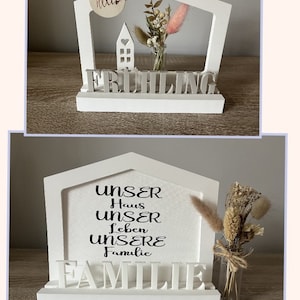 Könnte beinhalten: Ein weißes, hausförmiges Schild aus Holz mit dem Wort "FRÜHLING" in grauen Buchstaben. Das Schild hat ein kleines Fenster und eine kleine Vase mit getrockneten Blumen darin. Das Schild steht auf einer Holzoberfläche.