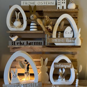 Könnte beinhalten: Ein Holzregal mit fünf weißen, eiförmigen Dekorationen mit Hasen und Ostergrüßen. Das Regal ist mit Trockenblumen, Holzhäusern und einem Schild mit der Aufschrift "Frohe Ostern" dekoriert.
