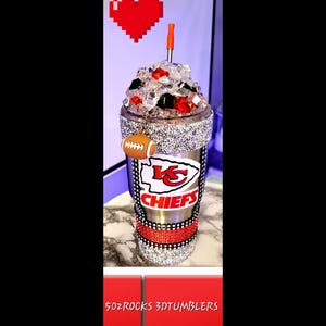 K&ouml;nnte beinhalten: Ein Edelstahlbecher mit einem rot-wei&szlig;-schwarzen Logo der Kansas City Chiefs. Der Becher ist mit Strasssteinen verziert und hat einen rot-wei&szlig; gestreiften Strohhalm. Der Becher ist mit Eis gef&uuml;llt und hat einen Fu&szlig;ball-Anh&auml;nger am Deckel.