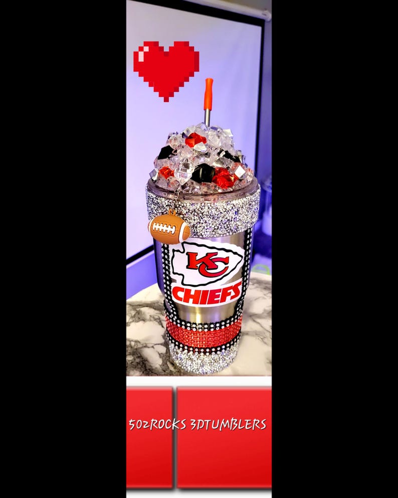 K&ouml;nnte beinhalten: Ein Edelstahlbecher mit einem rot-wei&szlig;-schwarzen Logo der Kansas City Chiefs. Der Becher ist mit Strasssteinen verziert und hat einen rot-wei&szlig; gestreiften Strohhalm. Der Becher ist mit Eis gef&uuml;llt und hat einen Fu&szlig;ball-Anh&auml;nger am Deckel.
