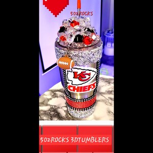 K&ouml;nnte beinhalten: Ein silberner und schwarzer Edelstahlbecher mit einem rot-wei&szlig;-schwarzen Logo der Kansas City Chiefs. Der Becher ist mit Strasssteinen verziert und hat einen durchsichtigen Deckel mit einem roten Strohhalm. Der Becher hat einen Fu&szlig;ball-Anh&auml;nger. Der Text "502 ROCKS 3DTUMBLERS" ist am unteren Rand des Bildes zu sehen.