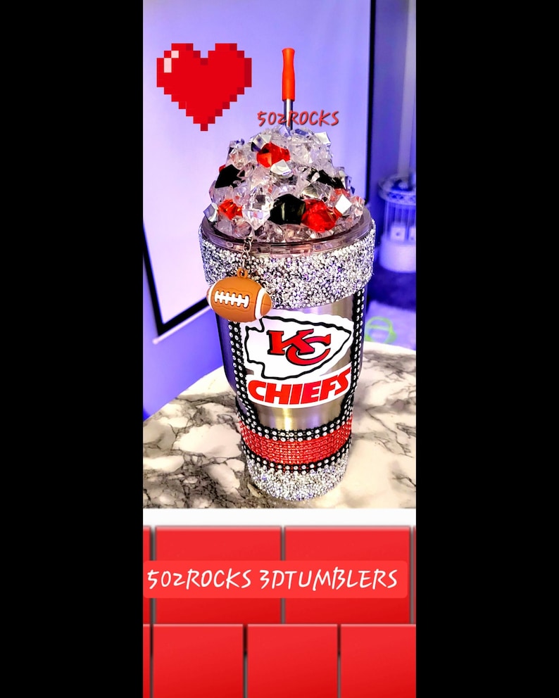 K&ouml;nnte beinhalten: Ein silberner und schwarzer Edelstahlbecher mit einem rot-wei&szlig;-schwarzen Logo der Kansas City Chiefs. Der Becher ist mit Strasssteinen verziert und hat einen durchsichtigen Deckel mit einem roten Strohhalm. Der Becher hat einen Fu&szlig;ball-Anh&auml;nger. Der Text "502 ROCKS 3DTUMBLERS" ist am unteren Rand des Bildes zu sehen.