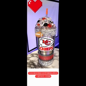 K&ouml;nnte beinhalten: Ein silberner und roter Chiefs-Becher mit Strasssteinen und einem Fu&szlig;ball-Anh&auml;nger. Der Becher ist mit Eis gef&uuml;llt und hat einen roten Strohhalm. Der Becher ist mit dem Logo der Kansas City Chiefs und dem Wort "Chiefs" verziert.