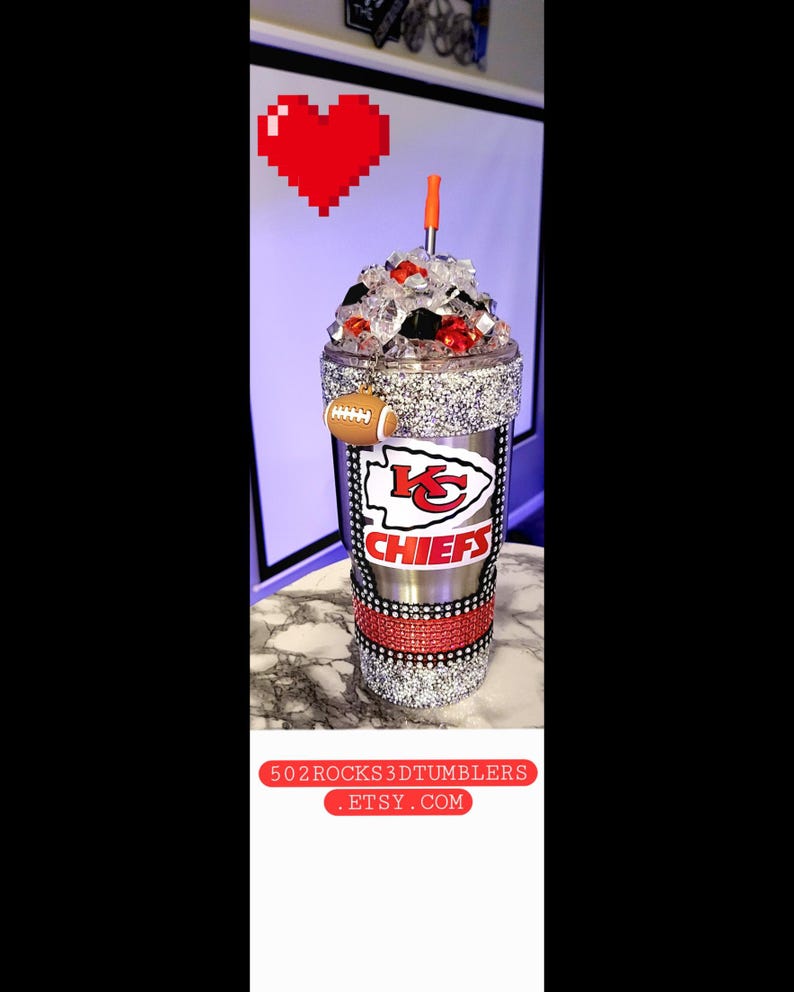 K&ouml;nnte beinhalten: Ein silberner und roter Chiefs-Becher mit Strasssteinen und einem Fu&szlig;ball-Anh&auml;nger. Der Becher ist mit Eis gef&uuml;llt und hat einen roten Strohhalm. Der Becher ist mit dem Logo der Kansas City Chiefs und dem Wort "Chiefs" verziert.