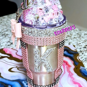May include: A stainless steel tumbler covered in pink and silver rhinestones with a large letter K in the center. The lid features a faux whipped cream topping with clear crystals, a pink bow, and a pink lipstick-shaped charm attached.