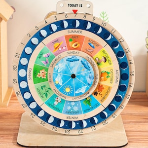 Może przedstawiać: Drewniane koło kalendarzowe z niebiesko-białym śledzeniem faz księżyca, kolorowym kalendarzem miesięcznym i kalendarzem dziennym z dniami tygodnia. Kalendarz zawiera również śledzenie pór roku z ilustracjami pór roku.