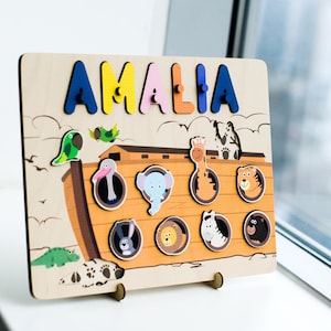 May include: Wooden puzzle board with the name "AMALIA" in colorful letters. Features illustrations of animals and Noah's Ark. Includes a parrot, elephant, giraffe, tiger, rabbit, lion, zebra, and bear. The board is supported by a wooden stand.