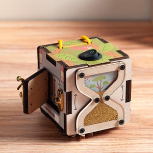 Puede incluir: Un cubo de actividades de madera con un interruptor basculante negro y un cordón amarillo en la parte superior. Un lado muestra un marco transparente en forma de reloj de arena con cuentas doradas, y otro lado tiene una puerta de madera abierta con pestillo.