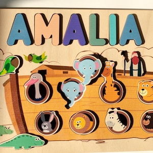 Puede incluir: Un rompecabezas de madera con el nombre AMALIA en letras coloridas sobre una ilustración del Arca de Noé. El tablero incluye recortes para varias figuras de animales, incluyendo elefantes, una jirafa, un tigre, un león, una cebra y un oso, junto a una figura de Noé.