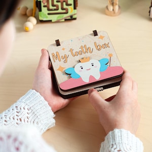 Op de afbeelding: Houten "My tooth box" met een cartoon tand ontwerp. De doos heeft een scharnierend deksel en heeft een tand met een kroon, blauwe vleugels en een roze basis. De tekst "My tooth box" staat boven de tand in een speels lettertype.