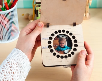 Puede incluir: Una caja de madera con una tapa con bisagras, que revela una foto de un niño pequeño con una camisa azul. La caja tiene un recorte circular con 20 círculos pequeños alrededor, y las fechas "05.02.19" y "04.02.19" están escritas en el lateral.