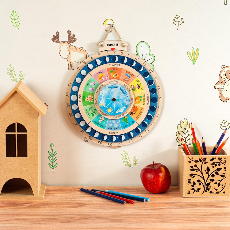 May include: A wooden educational calendar with a blue and white design, featuring the days of the week, months, and seasons. The calendar is hanging on a wall with decorative stickers. A wooden house, pencils, and an apple are on a wooden surface.