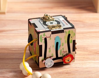 May include: A wooden activity cube featuring a top metal latch, a black rocker switch, and a yellow string with four wooden beads. The sides display printed illustrations of trees, a rhino, and a lion, with integrated maze tracks for sliding pieces.