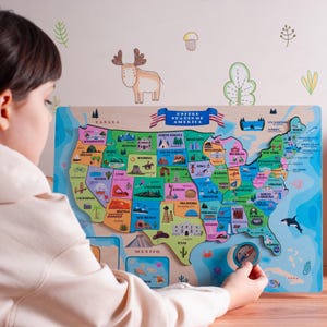 May include: A wooden United States map puzzle with colorful state outlines and illustrations. The puzzle includes the text "UNITED STATES OF AMERICA" and is being held by a person.