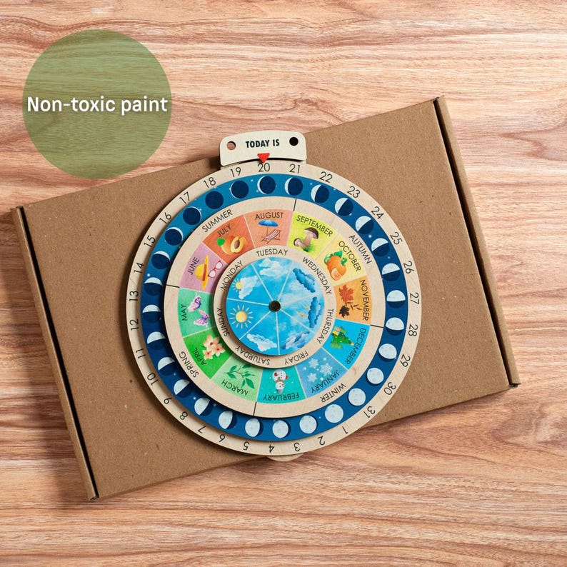 May include: Wooden educational calendar with a rotating dial showing months, seasons, and the phases of the moon. The calendar features colorful illustrations and the text "Non-toxic paint" is visible. The calendar is on top of a brown cardboard box.