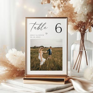May include: A gold-coloured framed table number sign. The sign displays the word "Table" in script, the number "6", a photo of a couple, the names "Emily & Kevin", and the date "08.26.20XX". The sign is on a gold stand.