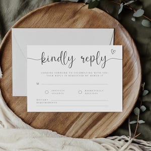 May include: A white RSVP card with black text that reads "kindly reply". The card has a small heart symbol and a section for guests to indicate whether they will be able to attend the event. The card also includes a section for dietary requirements.