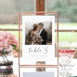 May include: A rose gold table number holder with a white card that says "table 3 Olivia & Thomas 16 August 20XX". The card has a photo of a couple in the background.
