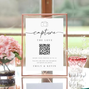 May include: A white sign with a black camera icon and the text "capture the love" in a cursive font. A black and white QR code is below the text. The sign also includes the text "Share your photos with us. Scan the code with your smartphone and upload your photos. Emily & Kevin."
