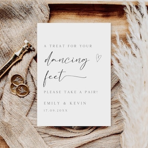 May include: A white card with black text that reads "A treat for your dancing feet. Please take a pair! Emily & Kevin 17.09.20XX". The card is on a wooden surface with a brown fabric and a white fluffy plant.