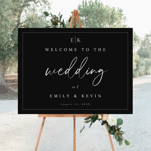 May include: Black wedding welcome sign with white text. The sign reads "E|K Welcome to the wedding of Emily & Kevin August 10, 20XX".