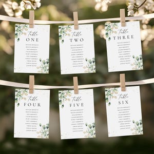 Può includere: Sei segnaposto bianchi con nomi, tenuti da clip di legno su un nastro. Ogni segnaposto ha un numero di tavolo e un elenco di nomi, con accenti di foglie verdi e dorate. Segnaposto per un matrimonio o evento.