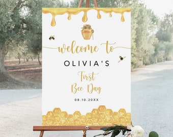 May include: White welcome sign with gold dripping honey, a honey jar, and bees. The sign says "Welcome to Olivia's First Bee Day 08.10.20XX". The bottom of the sign has a honeycomb pattern.