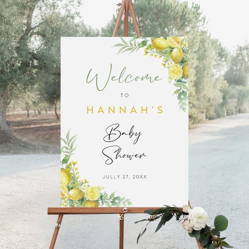 May include: Welcome to Hannah's Baby Shower sign with a lemon and greenery design. The sign is white with gold lettering and says "Welcome to Hannah's Baby Shower" and "July 27, 20XX".