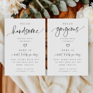 Puede incluir: Dos letreros de boda rectangulares con caligrafía elegante. Uno dice "Hello Handsome" y el otro "Hello Gorgeous". Ambos letreros incluyen el texto "Please help yourself" y "Here is a basket to help you along."
