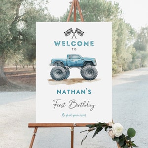May include: A white welcome sign with blue and black checkered flags, a blue monster truck, and the text "WELCOME TO NATHAN'S First Birthday So glad you're here!"