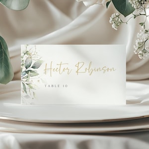 Könnte beinhalten: Eine weiße Tischkarte mit goldenem Blattdesign und goldener Schrift. Der Name "Hector Robinson" und "Tisch 10" sind auf die Karte gedruckt.