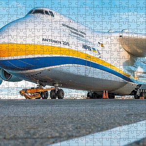 Pode incluir: Um grande avião de carga branco com listras azuis e amarelas. O avião está na pista com o trem de pouso abaixado. O avião está etiquetado como "ANTONOV 225" e "MPIB".