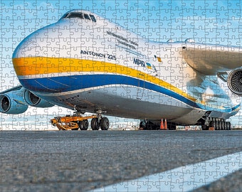 May include: A large, white cargo plane with blue and yellow stripes. The plane is on the tarmac with its landing gear down. The plane is labeled "ANTONOV 225" and "MPIB".