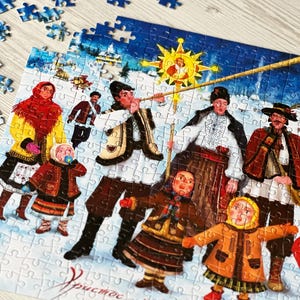 May include: A partially assembled jigsaw puzzle featuring a winter scene with figures in traditional clothing. The puzzle depicts a festive gathering with a star and text in a foreign language. Blue puzzle pieces are scattered around.