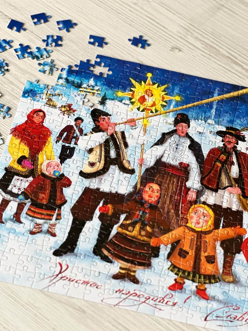May include: A partially assembled jigsaw puzzle featuring a winter scene with figures in traditional clothing. The puzzle depicts a festive gathering with a star and text in a foreign language. Blue puzzle pieces are scattered around.