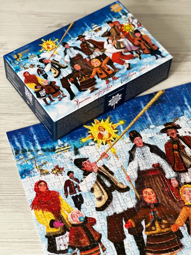 May include: A jigsaw puzzle with a winter scene featuring people in traditional clothing, a sun, and text. The puzzle box is blue with the same image. The puzzle pieces are white with a detailed illustration.