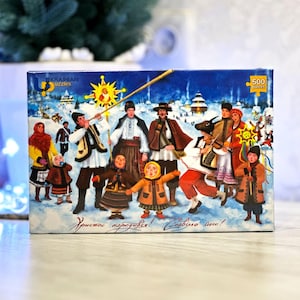 May include: A 500-piece jigsaw puzzle featuring a winter scene with people in traditional Ukrainian clothing. The box has the text "KRAIMAN puzzles" and a greeting in Ukrainian. The artwork depicts a festive holiday celebration.