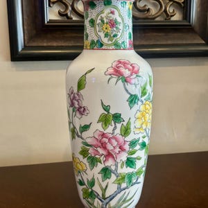 May include: A white ceramic vase with a floral design. The vase features pink, yellow, and purple flowers with green leaves. The top and base have decorative patterns. The vase is approximately 12 inches tall.