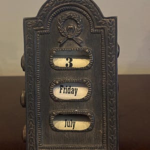 Peut inclure: Un calendrier de bureau perpétuel en métal sombre avec un sommet arqué et un détail de couronne de laurier en relief. L'avant présente trois fenêtres horizontales affichant le chiffre 3, le jour Friday et le mois July sur des rouleaux de papier blanc cassé.