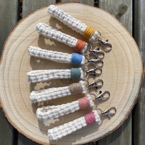 May include: A selection of handmade macrame keychains. Each keychain has a white woven cord with a coloured accent band in yellow, orange, blue, green, tan, pink, and rose. They feature silver-coloured lobster claw clasps.
