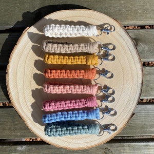 May include: A selection of macrame wrist keychains in shades of white, beige, mustard, orange, pink, blue, and teal. Each keychain has a woven macrame strap and a silver-coloured clasp for attaching keys. The keychains are displayed on a wooden surface.