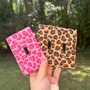 May include: Two rectangular light switch plates featuring animal print patterns. One plate has a bright pink and magenta leopard spot design for a single toggle switch. The other plate is a double toggle switch cover with a tan and brown leopard print.