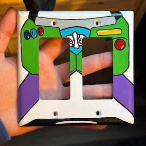 May include: A double light switch wall plate featuring a hand-painted design inspired by a space ranger suit. The plate is primarily white with green, purple, blue, and red accents, including a central blue emblem and two rectangular cutouts.