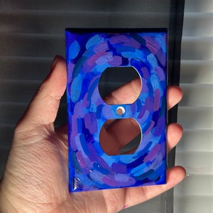 May include: A hand-painted, decorative electrical outlet cover. The cover is painted with a swirling pattern of blues and purples. The cover has two circular openings for the electrical outlets and a small hole for a screw.