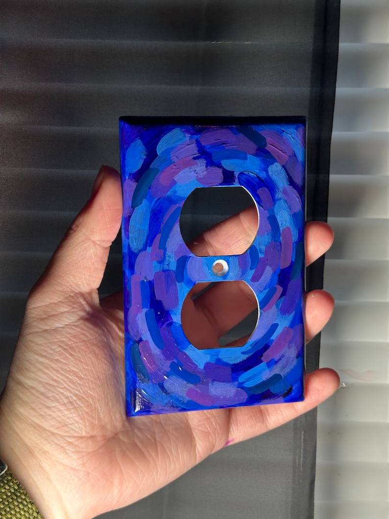 May include: A hand-painted, decorative electrical outlet cover. The cover is painted with a swirling pattern of blues and purples. The cover has two circular openings for the electrical outlets and a small hole for a screw.