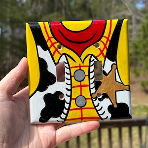 May include: A square light switch cover plate featuring a hand-painted design of a yellow shirt with a red collar, black and white cow print vest pattern, and a gold sheriff star badge. The plate has two rectangular cutouts and two screw holes.