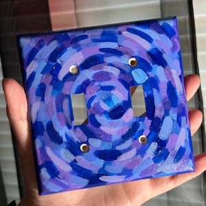 May include: A light switch cover painted with a swirling pattern of blue, purple, and white paint. The cover has two toggle switches and four screw holes.