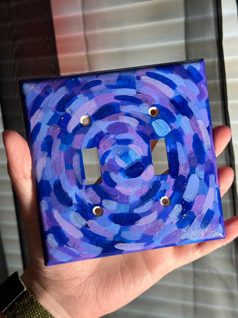 May include: Hand-painted double light switch cover with a swirling design in shades of blue and purple. The cover has a black border and two switch openings with brass-colored screw holes. The artwork features brushstrokes creating a textured appearance.