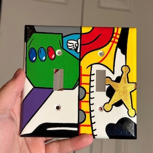 May include: A hand-painted light switch cover featuring Buzz Lightyear and Sheriff Woody from Toy Story. The left side is green, purple, and white, while the right side is yellow, red, and black. The cover has two switch openings and two screw holes.